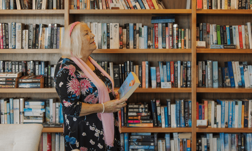 An older woman with white hair, wearing a floral dress and pink scarf, stands in front of a large bookshelf filled with books, holding an open book and looking up thoughtfully.
