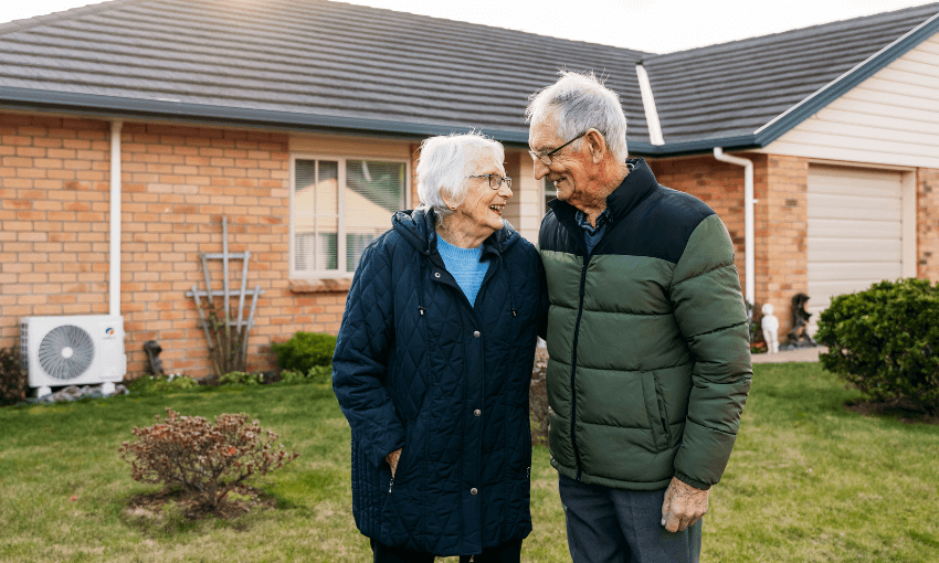 An elderly couple stands smiling and embracing outside a brick house, both wearing winter jackets. The lawn is green, and the house features a garage and a window.