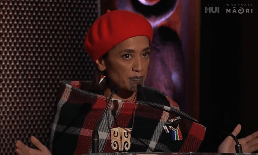 Oriini Kaipara speaks from behind a podium with her hands flayed out.