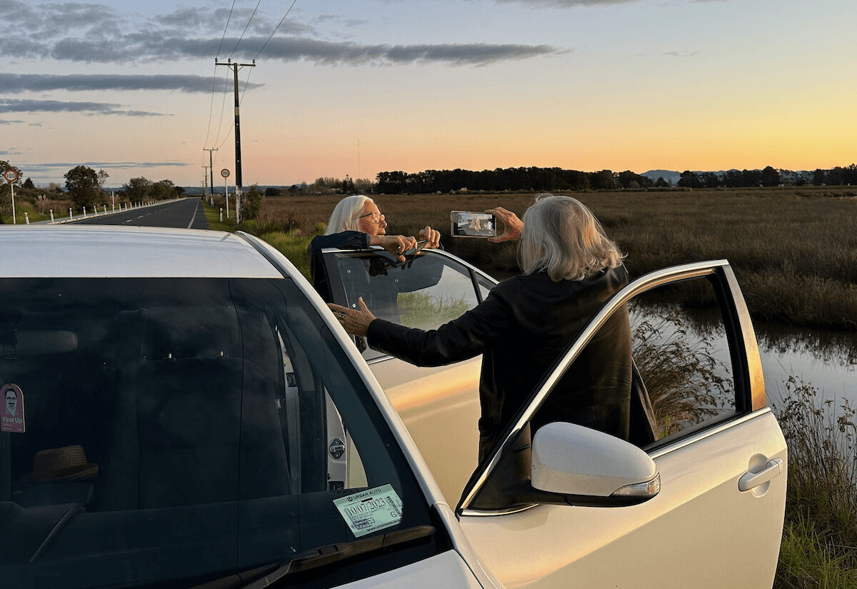 two white haired ladies by a white car looking out at the sunset. One is filming the other on a phone