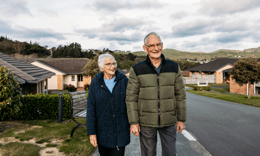 An elderly couple smiles while walking hand in hand outside in a suburban neighborhood, with houses, greenery, and hills in the background on a partly cloudy day.