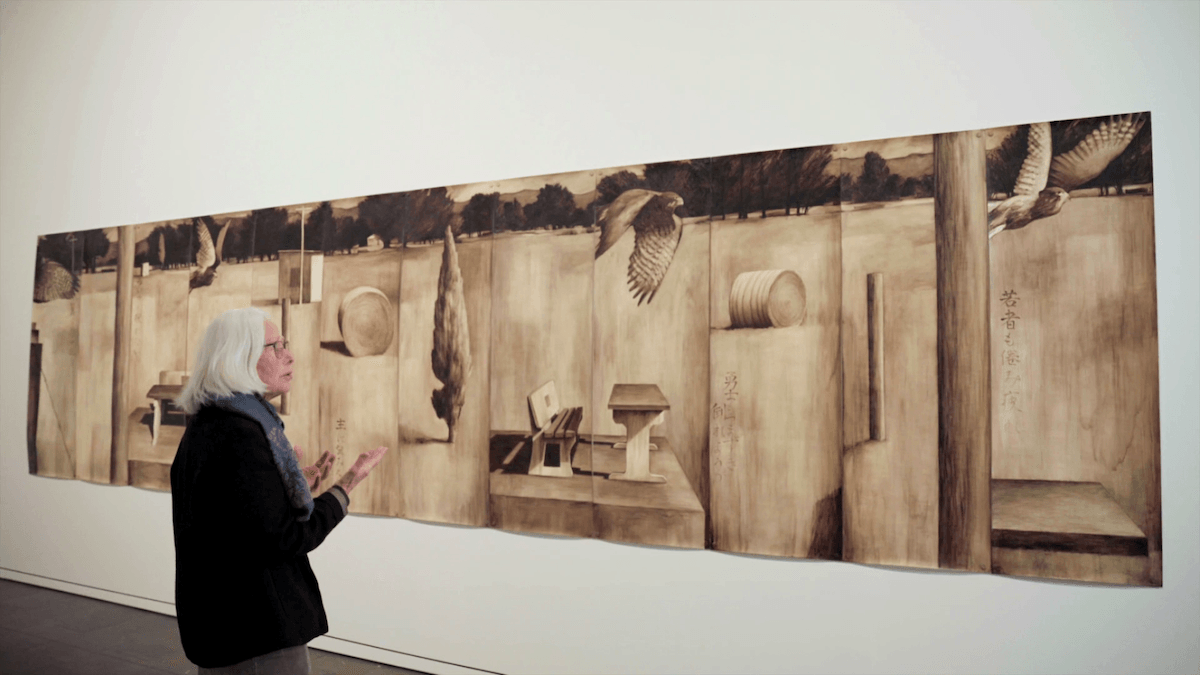 A woman standing in front of a large multi-panel monochrome painting of a landscape. it is painted on the back of wallpaper