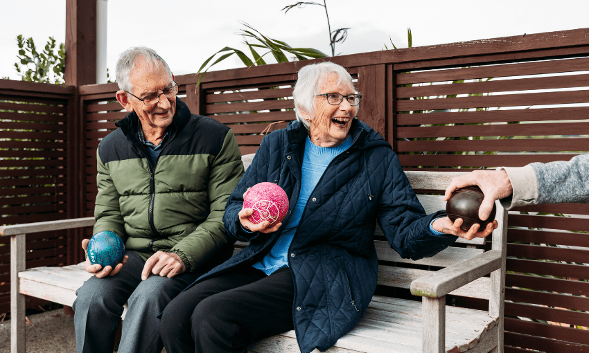 Two elderly people sit on a bench outdoors, smiling and holding colorful lawn bowls. One person hands a black bowl to the woman, who is laughing and wearing a blue quilted jacket.