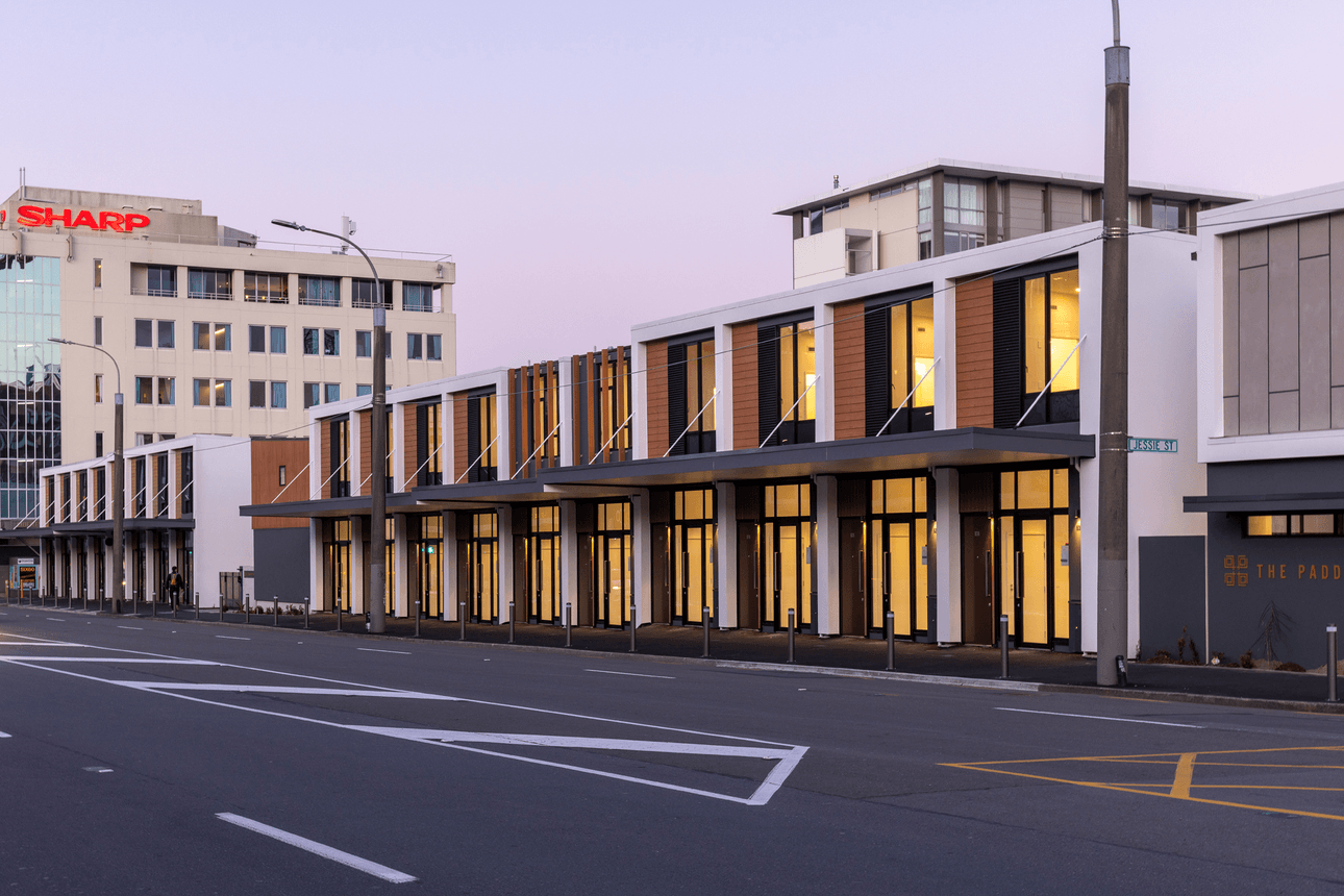 Modern row of two-story townhouses with large windows and balconies, illuminated warmly in the early evening. The street in front is empty, with a commercial building visible in the background.