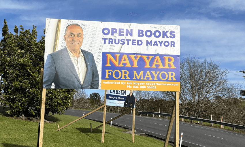 A large roadside campaign sign features a smiling man in a suit with text reading "Open Books Trusted Mayor, NAYYAR FOR MAYOR." There is a smaller Larsen campaign sign below it, and trees and a road are visible in the background.