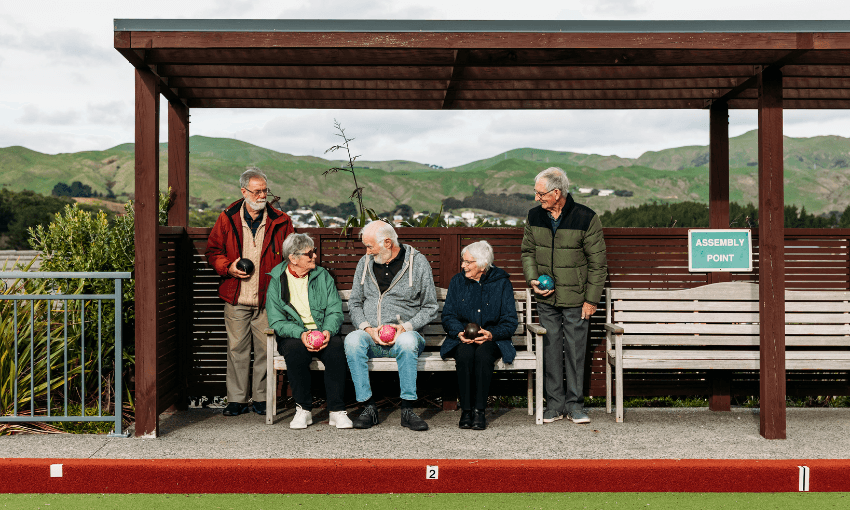 Five seniors, three seated and two standing, hold lawn bowls under a wooden shelter by a green lawn. Hills and houses are visible in the background. A sign reads "Assembly Point.