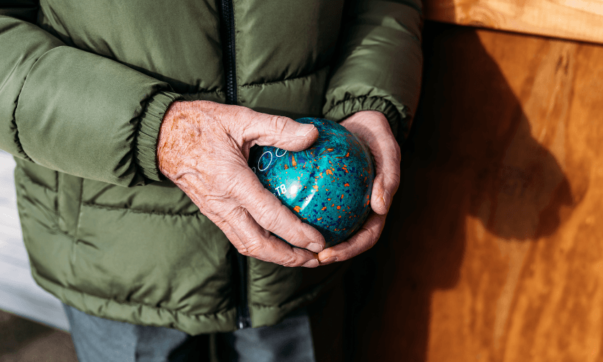 An older person wearing a green jacket holds a round, blue, speckled glass object in their hands, standing next to a wooden surface in natural light.