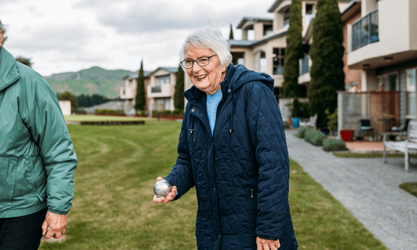 An elderly woman in a navy coat smiles while holding a metal ball outdoors, standing on a grassy area near modern houses. Another person in a green jacket is partially visible beside her.