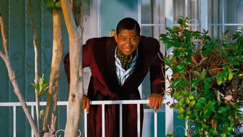 A man wearing a dark robe and patterned scarf leans over a white railing on a porch, surrounded by plants and greenery, with a house and window in the background.