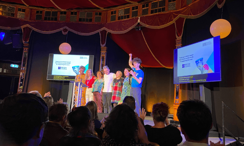Six poets on stage with two screens on either side of them. The venue is fancy with red velvet drapes.