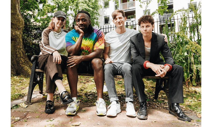 A photo of the band Bloc Party - all four band members are sitting on a park bench and smiling. It is summery.