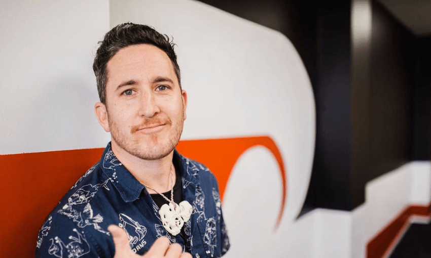 A man with short dark hair and a trimmed beard stands indoors, wearing a dark patterned shirt and a carved pendant necklace. He is smiling slightly and giving a shaka hand gesture, with a modern red, white, and black wall behind him.