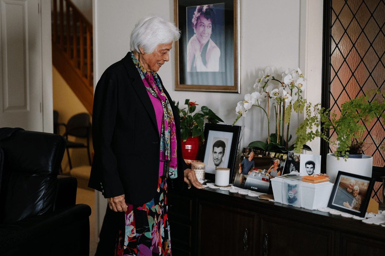 an older woman with white hair stands next to a side table looking at framed photos. she wears a colourful long dress and a black blazer