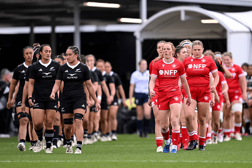 two rugby teams, one in red the other in black, walk onto a grass field in single file