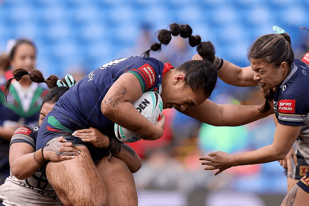 a woman rugby player is tackled by two opponent players, one around the legs and one in front