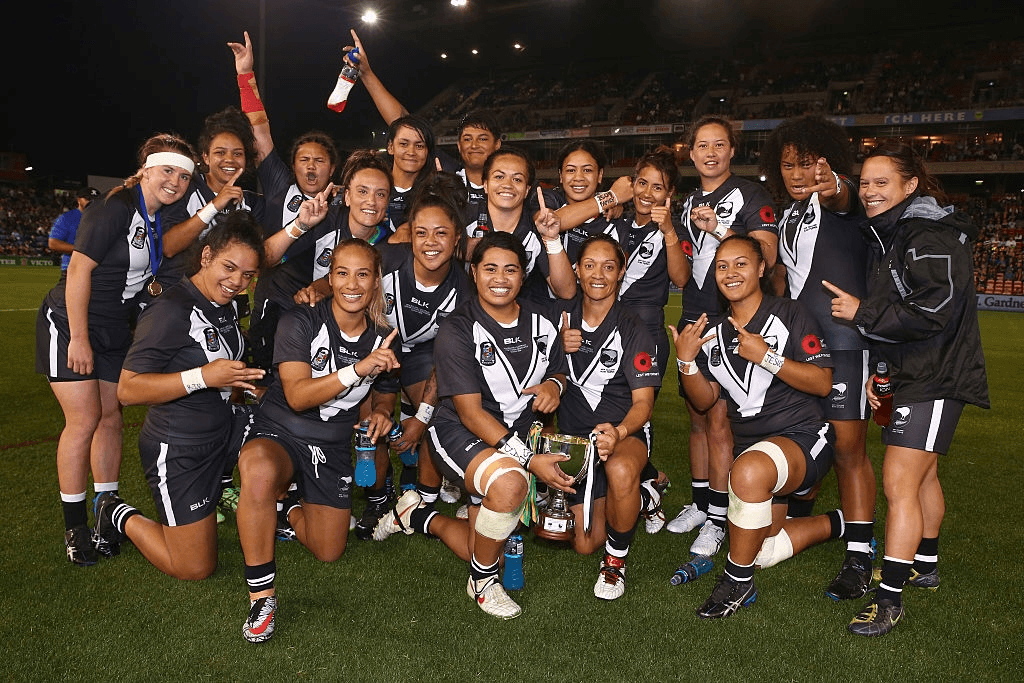 a team of women rugby league players in black uniforms celebrate to the camera