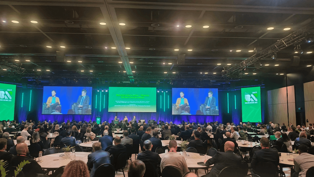 A large conference room filled with white tables and people sitting around them, watching three people speak on stage at the front