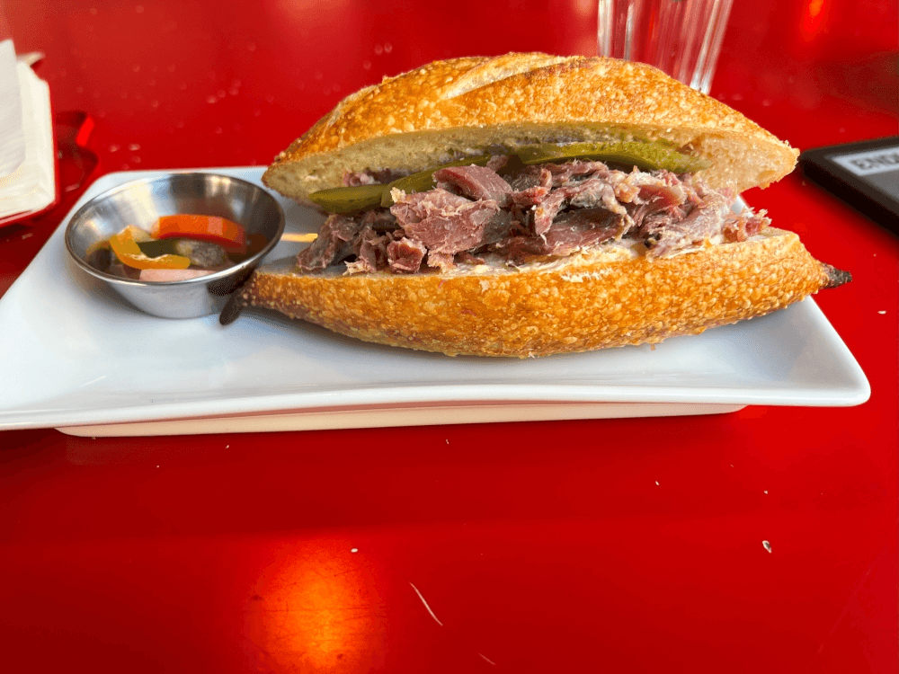 A sandwich with shredded meat and pickles on crusty bread sits on a white plate, next to a small dish of mixed pickled vegetables, on a bright red table. A glass of water and napkins are in the background.