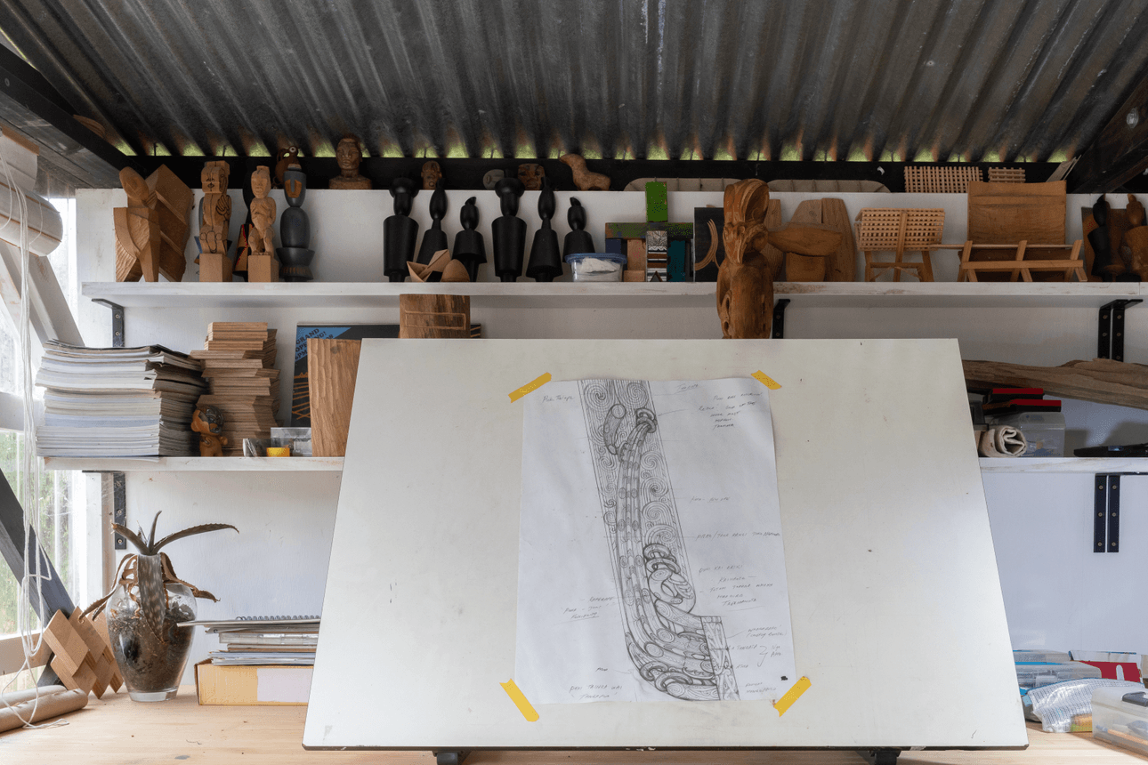 A sketch of a whakairo design is pinned to a tilted drawing board inside Leon Kipa’s studio. Shelves above hold carved figurines, timber samples, and small sculptural works in progress.