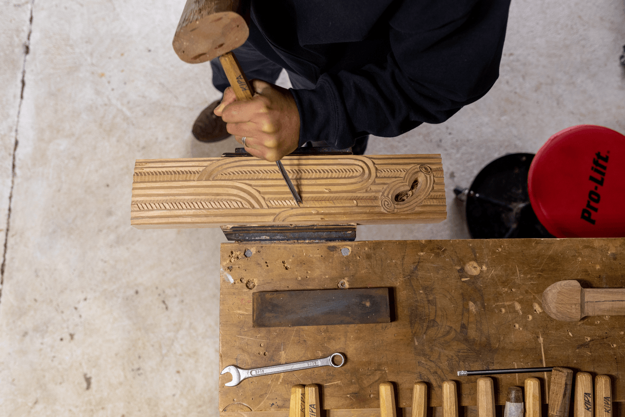A top-down view of Leon Kipa carving intricate grooves and motifs into a piece of timber secured to a workbench. His tools, mallet, and a red workshop stool are visible beside him.