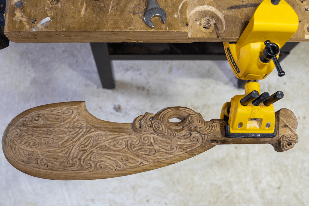 A carved wooden patu lies secured in a yellow clamp on Leon Kipa’s workbench. Its blade is adorned with swirling motifs, surrounded by wood shavings and hand tools.