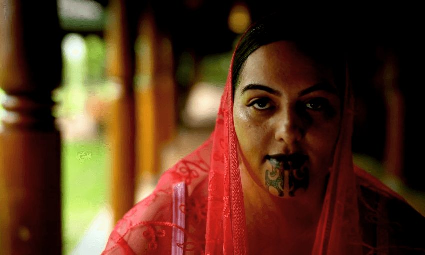 A woman draped in a sheer red veil looks toward the camera, with traditional black chin tattoos visible on her lower face. Wooden columns and greenery are blurred in the background.