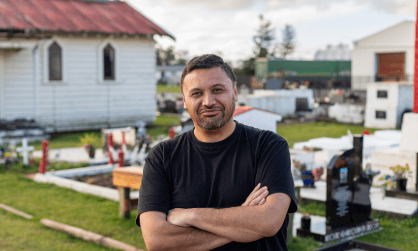 A man with short dark hair and a beard stands with his arms crossed in a cemetery, with graves and a white wooden building visible in the background.