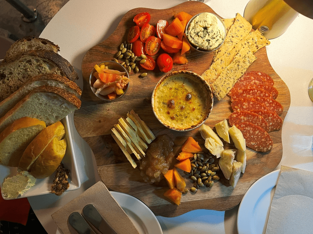 A wooden platter with sliced bread, crackers, sliced cured meats, cheese, artichokes, cherry tomatoes, pickled vegetables, pumpkin seeds, spreads, and dip, set on a white table with plates and cutlery nearby.