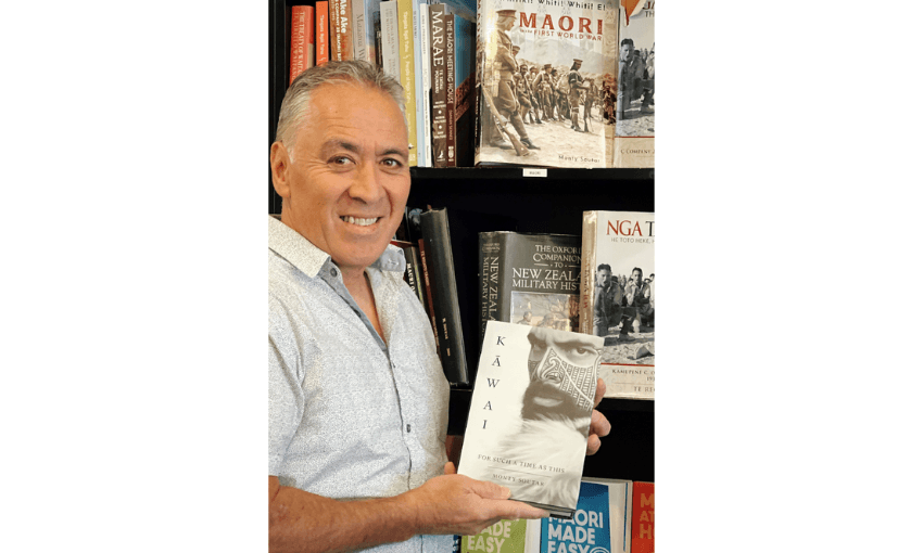 Author Monty Soutar holding his novel Kāwai and standing in front of bookshelves. Monty is similing.