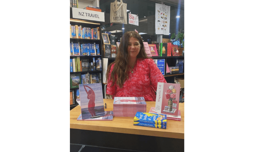 A photo of Nina Mingya Powels who has long brown hair and is wearing a red-pink dress. She is standing at a shop counter and has her books around her.