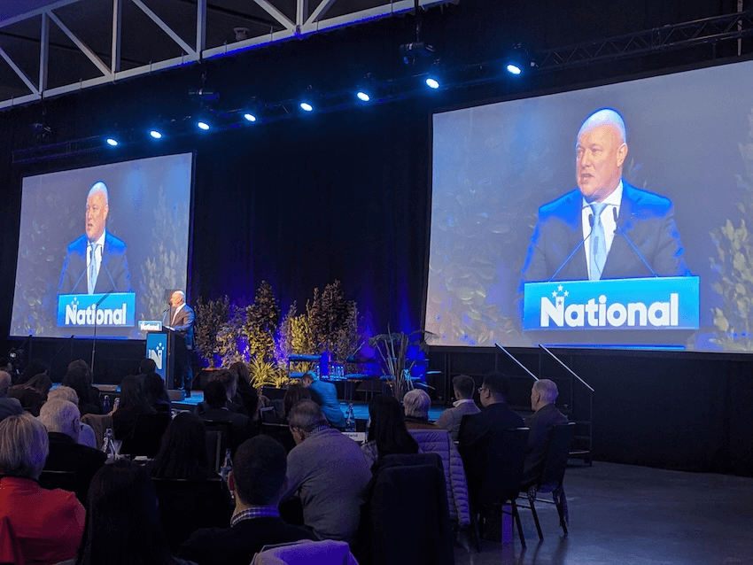 a blue toned ohoto of some tables and people looking at Christopher Luxon, a bald white man, wearing a suit with his face projected behind him