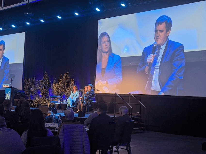 indoor conference room with blue lights; nicola Willis, in a powder blue suit, looks at Chris Bishop, who is holding a microphone