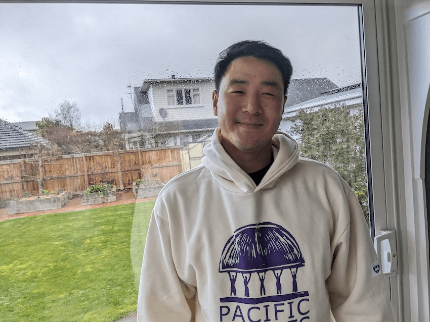 a young man in a pacific studies hoody, smiling at the camera with a rainy garden outside