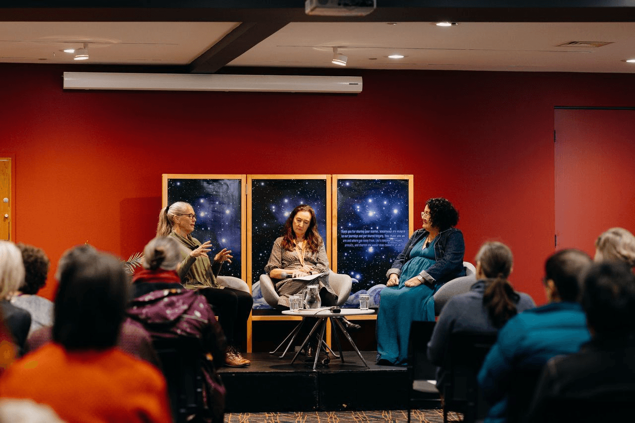 Three people sit on a stage in front of an audience, engaged in a panel discussion. Behind them are posters with starry night images. The stage and walls are red, and the room is warmly lit.
