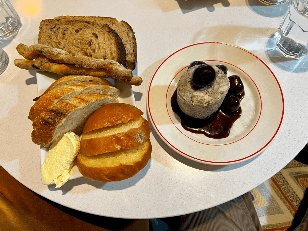 A plate with assorted breads and butter sits beside a dessert topped with dark cherries and sauce on a small round table, with glasses of water in the background.