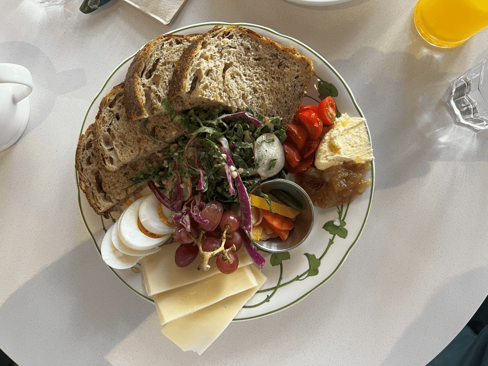 A plate with slices of multigrain bread, hard-boiled egg, cheese, grapes, salad greens, cherry tomatoes, pickles, butter, and jam, photographed from above on a white table.