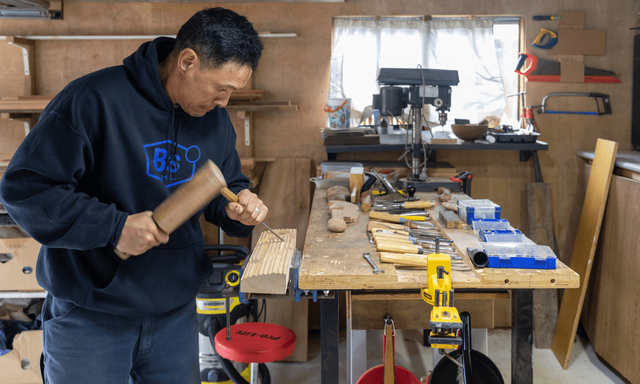 A man in a workshop uses a wooden mallet and chisel on a piece of wood at a workbench covered with tools, with woodworking equipment and supplies visible in the background.