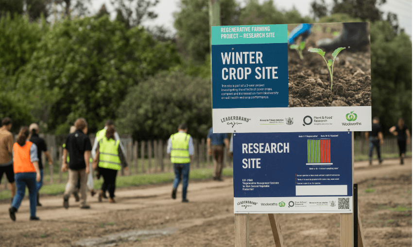A large sign for a winter crop and research site stands in the foreground, with people in high-visibility vests walking along a dirt path in a farm or research field in the background.