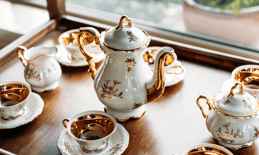A porcelain tea set with gold accents, including a teapot, cups, saucers, a creamer, and a sugar bowl, is arranged neatly on a wooden tray near a window.