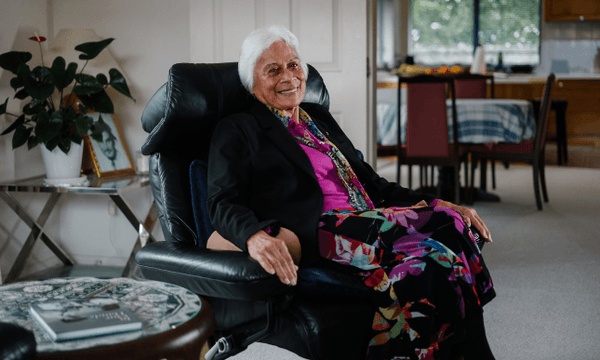 a woman with brown skin and white hair sits in an armchair at home, smiling at the camera