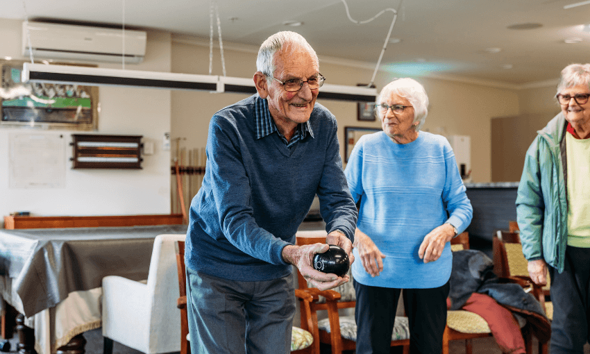 An elderly man smiles while holding a black bowling ball indoors, as two elderly women watch and smile beside him. The group appears to be enjoying a casual game in a community room.