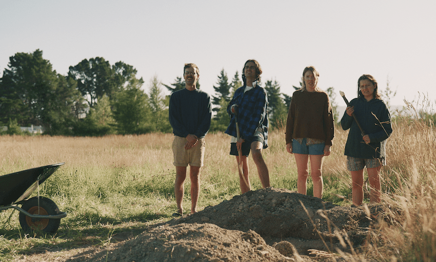 four people stand next to a freshly dug hole with an empty field behind them and a wheelbarrow beside them