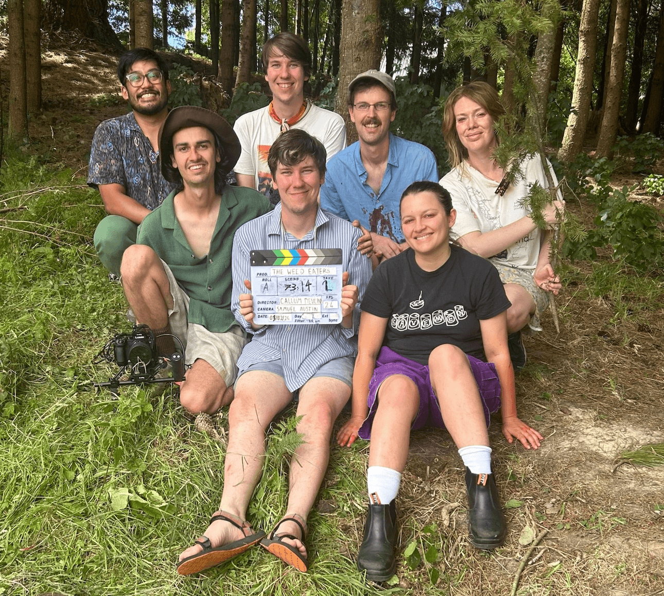 A group of people sit for a group photo on a bank with a sunny forest behind them