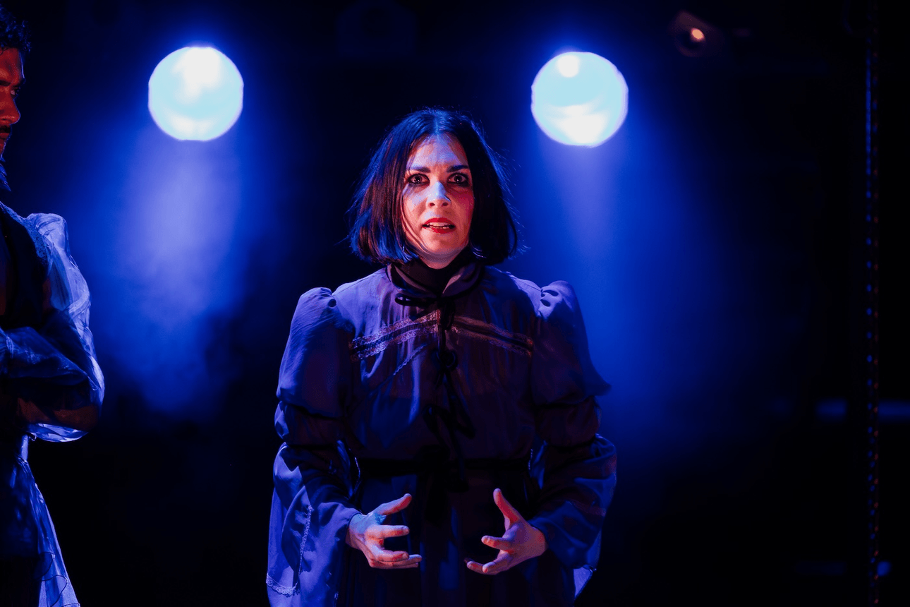 A woman with dark hair parted down the middle and wearing a dark purple dress stands centre stage