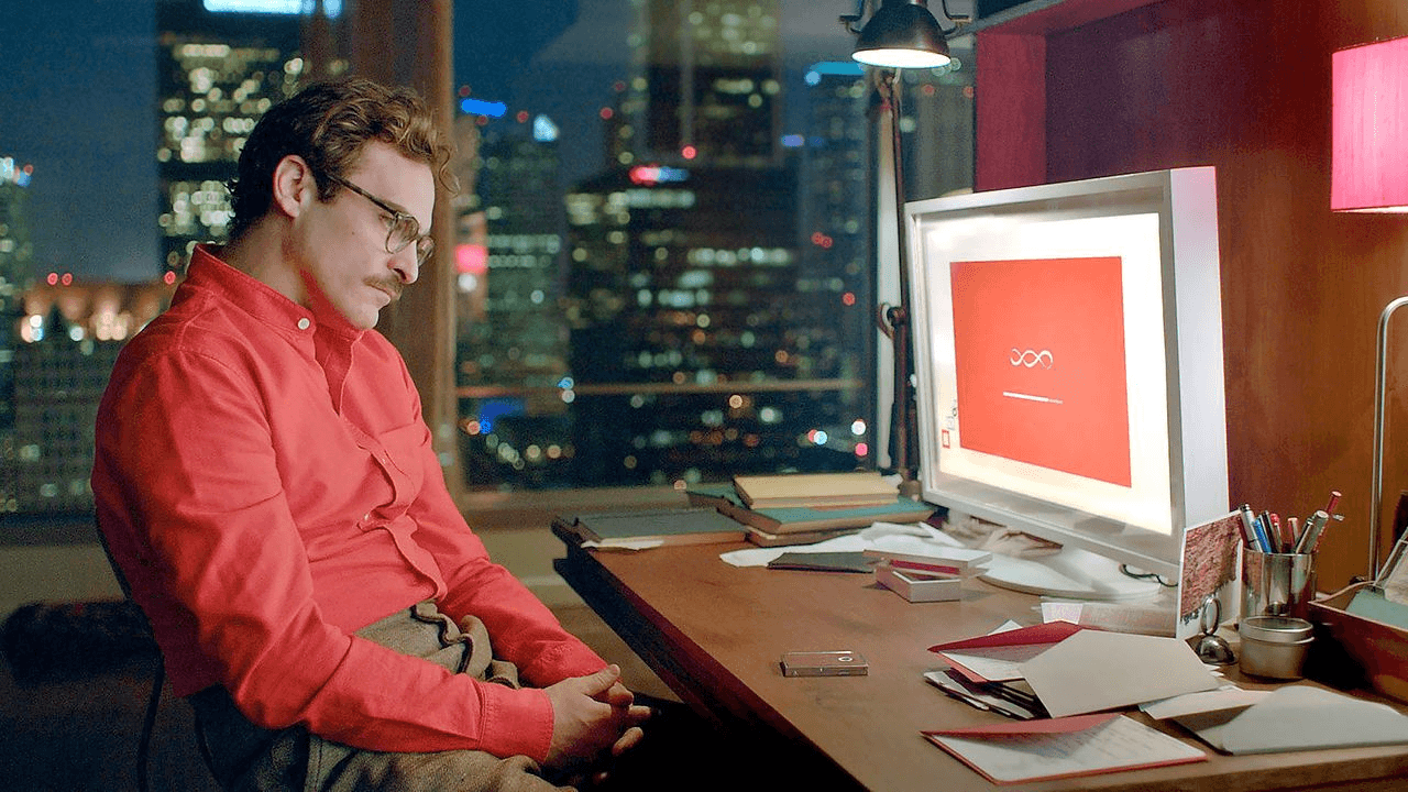 a man (joaqin phoenix) sits at a computer wearing a red shirt. there is a city view out the window behind him
