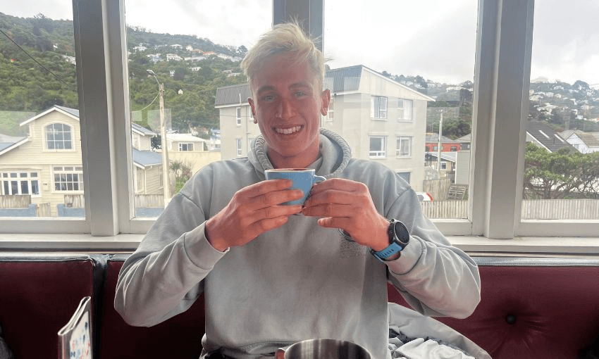 Luke Kuggeleijn smiles and holds a cup of coffee at Maranui Cafe in Lyall Bay.