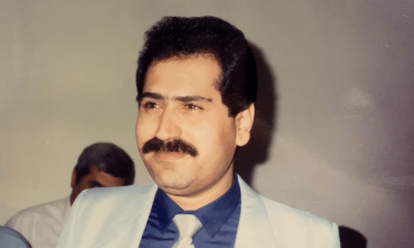 a man with dark hair and a dark moustache smiles off camera wearing a beige suit and blue shirt