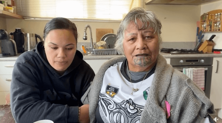 two women sit at a kitchen table. one is older and has a moko kaue and looks distressed. the other is younger and wearing a black hoodie