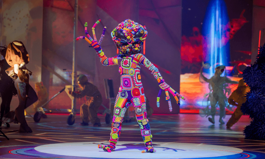 A performer in a colorful, crocheted costume with a large, multicolored headpiece and outstretched fingers stands on stage under bright lights, with people and abstract set pieces in the background.