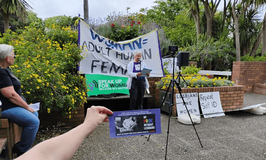 An older woman speaks in front of a "woman = adult human female" sign, while a separate hand holds up a "no pride without lesbians" sign.